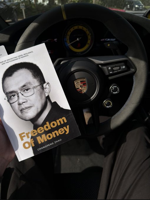 A person holds Changpeng Zhao's "Freedom of Money" book in a Porsche, fitting the @cz_binance tag as a nod to the Binance founder's autobiography about resilience.
