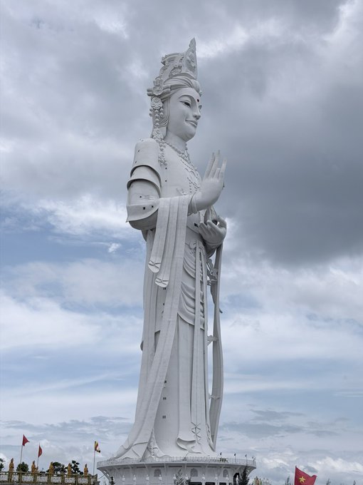 A towering white statue of a standing Buddhist deity (Guanyin) with ornate robes, an elaborate crown, and hands raised in prayer, set against a cloudy sky.