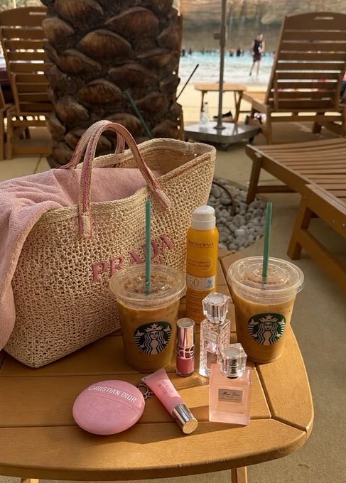 A poolside table scene featuring a Prada straw tote bag, two Starbucks iced coffees, sunscreen, perfume bottles, and a Christian Dior compact.