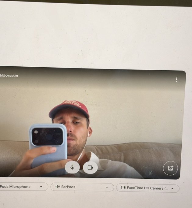 A man wearing a red cap and white earphones takes a mirror selfie with his smartphone during a video call on his computer, with input options showing 'Pods Microphone', 'EarPods', and 'FaceTime HD Camera'.
