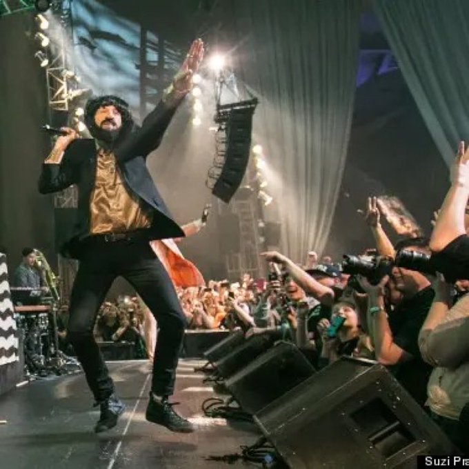 A bearded male performer in a black blazer and gold shirt stands on a stage with one hand raised, facing a crowd of fans holding up phones to record, credited to "Suzi P".