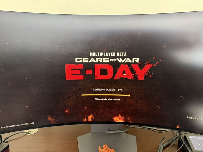 A curved monitor displaying the 'Gears of War: E-Day Multiplayer Beta' loading screen at 89% compiling shaders.