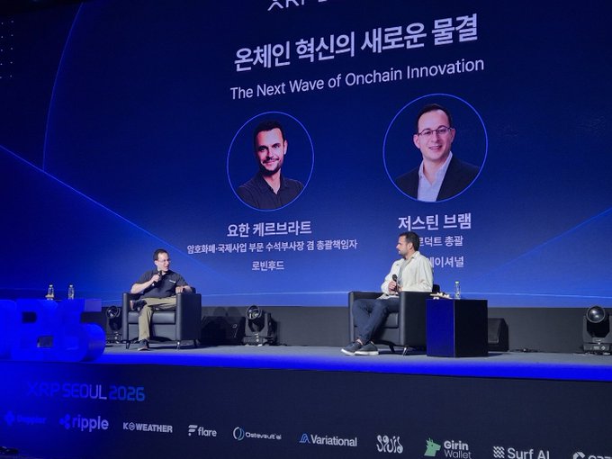 Two men are seated on stage at the XRP Seoul 2026 conference, discussing "The Next Wave of Onchain Innovation" as displayed on the large screen behind them. The screen features headshots of YoHan KeroBrant (Co-Founder of RootNode) and Justin Bram (Asia Strategic Lead at Onafriat), while the event is sponsored by Ripple, Weather, Flare, Onafriat, Variational, Solo, Griin Wallet, and Surf AI.