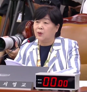 A woman in a striped blazer speaks into a microphone at a desk, with a nameplate reading '시영교' and a digital timer displaying '00:00'.