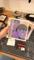 A slightly blurry photo of items laid out on a table, featuring a purple character card or figurine, what appears to be a Nintendo Switch console, and other small objects.