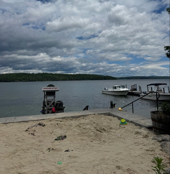 The image shows a beach scene with sand, water, boats, and trees. There are two boats docked at the shoreline, one of which has an "L" sign on top indicating that it's a marina or boatyard. The sky is cloudy, suggesting that it might be a cool day. In the foreground, there is a pile of sand with various beach toys scattered around, including a green bucket and shovel. A person can also be seen in the image, possibly enjoying their time at the beach.