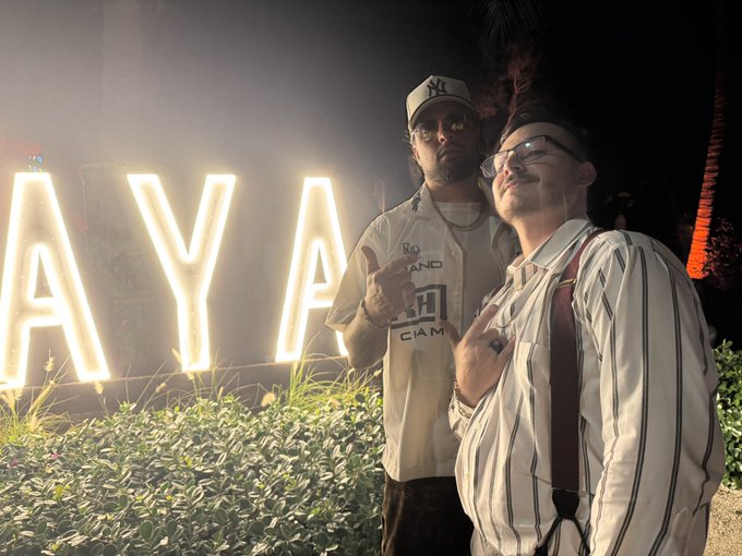 Two men pose at night in front of large, illuminated letters spelling 'AYA', with one wearing a New York Yankees cap and the other wearing glasses and suspenders.