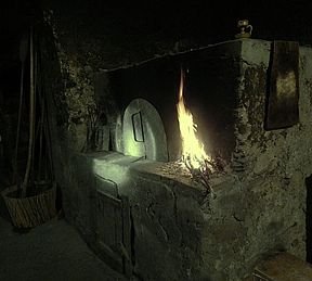 A dimly lit, atmospheric scene shows a rustic hearth or oven with a fire burning brightly inside its arched opening, casting light on the surrounding dark stone and a wooden broom leaning nearby.