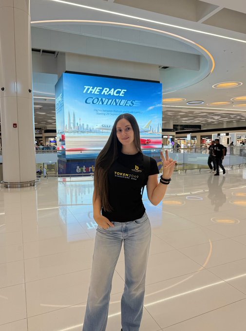 A woman wearing a black "Token2049 Singapore 2024" t-shirt poses with a peace sign in front of a "THE RACE CONTINUES" digital display at an airport, en route to Singapore for the Token2049 crypto conference with BenX Capital.