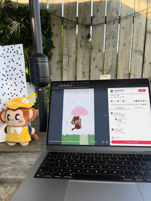 An outdoor wooden table scene with a laptop displaying a cartoon character holding a pink umbrella, alongside a monkey plushie in a yellow raincoat, with a pole and wooden fence in the background.