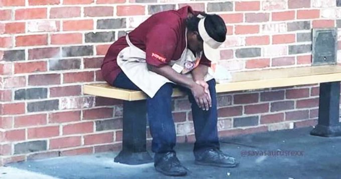 A pizza delivery worker in a red Papa John's uniform slumps forward on a wooden bench against a brick wall, appearing exhausted or dejected. Watermark: @savasaurusrex.