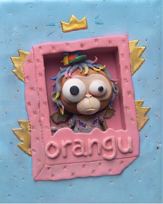 A claymation-style image of a small character with wild, multicolored hair, big googly eyes, and a small hat, framed within a pink border featuring a gold crown, set against a light blue textured background with gold accents and the word 'orangu' at the bottom.