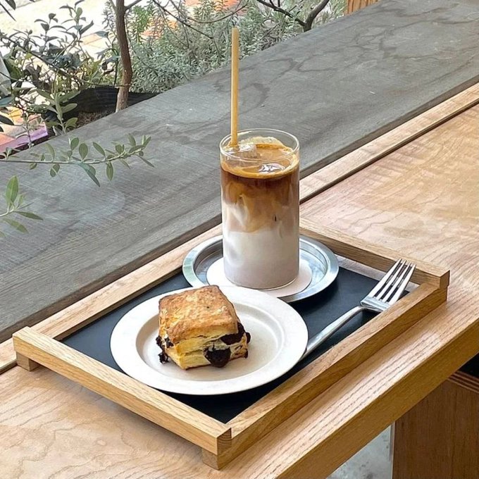 A wooden tray on a windowsill holding an iced coffee in a glass, a piece of cake on a plate, and a fork.