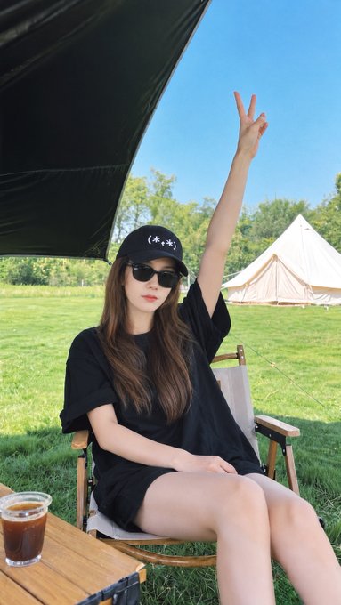 A young woman wearing a black t-shirt, black cap with a smiley face, and sunglasses sits in a camping chair on grass, raising one arm in a peace sign, with a glass of iced coffee nearby.