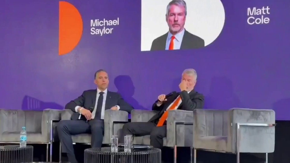 The image shows two men sitting in chairs at a conference room table, facing each other as they engage in conversation. The background features a large screen displaying an image of Michael Saylor, who appears confident about his Ethereum investment. The text on the screen reads "Michael Saylor - Confidence in Ethereum has collapsed. Ethereum now faces competition from Solana, BNB, Hyperliquid, and other L2s." This suggests that the men are discussing the current state of the cryptocurrency market and its impact on various companies.