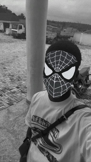 A black and white selfie shows a person wearing a Spider-Man mask and a graphic t-shirt, standing in an outdoor area with trucks and a motorcycle in the background.