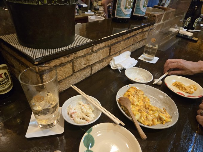 A bar counter with plates of Japanese food including tamagoyaki, a small bowl with chopsticks, empty glasses, sake bottles, and a person's hand reaching for a dish.