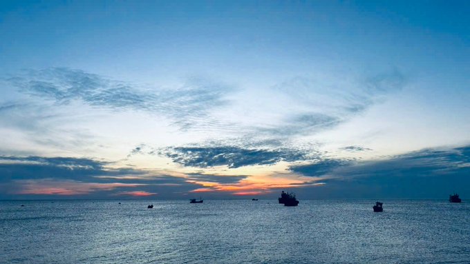 A serene ocean scene at dusk with several ships silhouetted on the horizon beneath a colorful sky of blues, oranges, and scattered clouds.