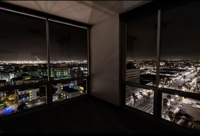 A view from inside a high-rise apartment at night, looking out through two corner windows at a sprawling city skyline illuminated with countless lights.