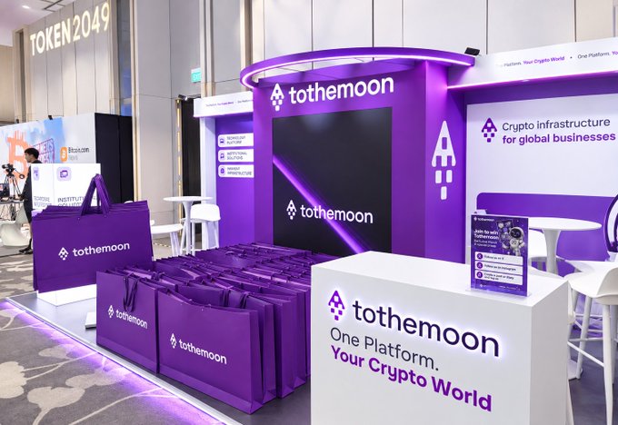 A purple-branded exhibition booth for 'tothemoon' at a TOKEN2049 event, featuring the tagline 'One Platform. Your Crypto World' and 'Crypto infrastructure for global businesses,' with branded tote bags displayed in front.