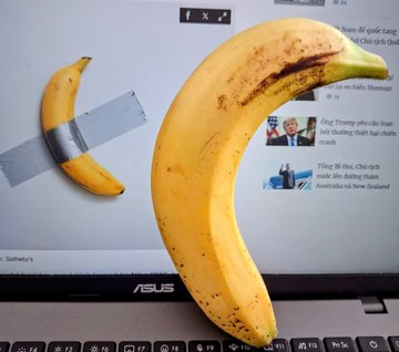 A real-life photo showing a banana taped to a laptop screen displaying a Sotheby's auction page, referencing the famous 'Comedian' artwork by Maurizio Cattelan.