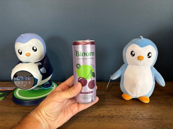 A hand holds a slim can of "Bloom" sparkling energy drink (Cherry Lime flavor) in front of two penguin plushies—one with a digital clock showing "09:36" and another holding a small object—lining up perfectly with the playful "GM to everyone addicted to hope" greeting, as the penguin companions and bubbly beverage capture a cheerful morning energy.