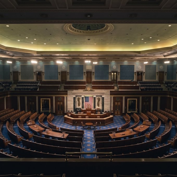 An empty interior view of a grand legislative chamber, likely the U.S. House of Representatives, featuring rows of curved wooden desks, blue carpeting, and an American flag at the front.