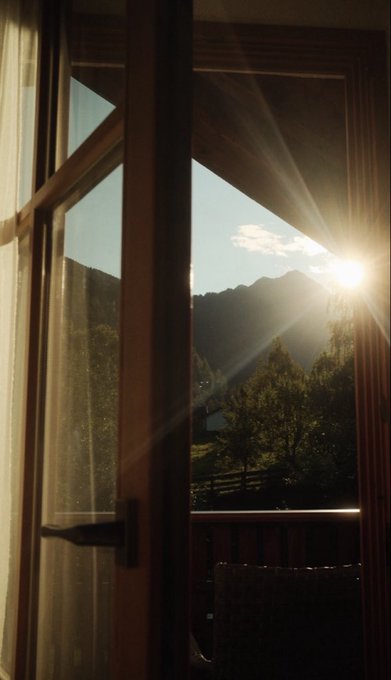 A sunburst streams through a partially open wooden balcony door, casting long rays across the frame with majestic mountains and lush trees visible beyond. The view captures a serene alpine escape, hinting at a tranquil retreat that would make any lucky winner of the 1,000 #Night giveaway eager to claim this breathtaking destination.