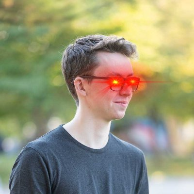 A young man with brown hair poses outdoors wearing the "matt black" Hack sunglasses, which appear to be glowing red—fitting the post's reference to those glasses. The eye-catching visual matches the caption's theme of "attack" and complements the accompanying crypto/tech commentary.