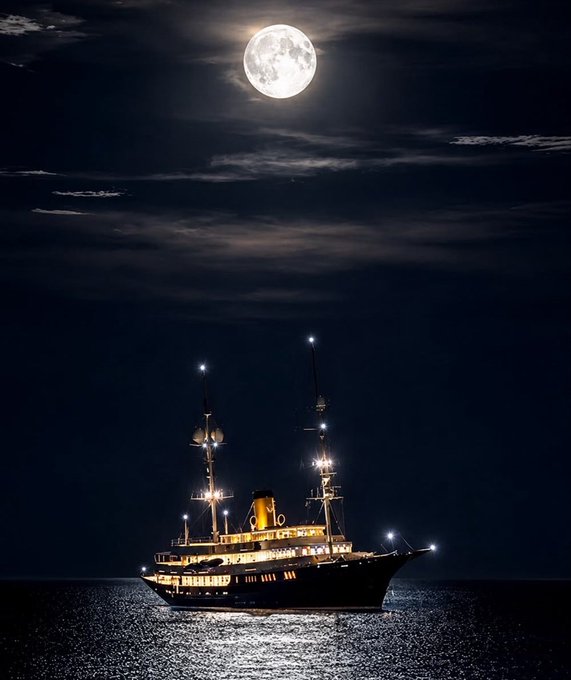 A large illuminated ship floats on calm dark waters under a bright full moon in a partly cloudy night sky, with the ship's warm lights reflecting on the sea's surface.