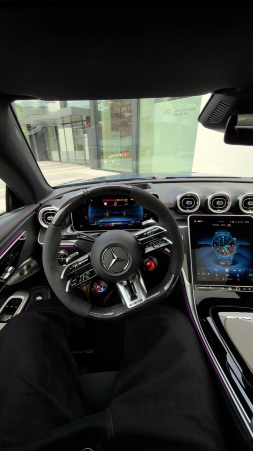 The image shows the interior of a Mercedes-AMG vehicle, featuring a black leather steering wheel with the Mercedes logo, dual digital displays, and AMG badging, with the "AMG" text visible on the center console.

The caption fits the post perfectly, as it suggests the user is flaunting a high-end Mercedes-AMG car, likely teasing followers with the opportunity for an in-person reveal or test drive experience.