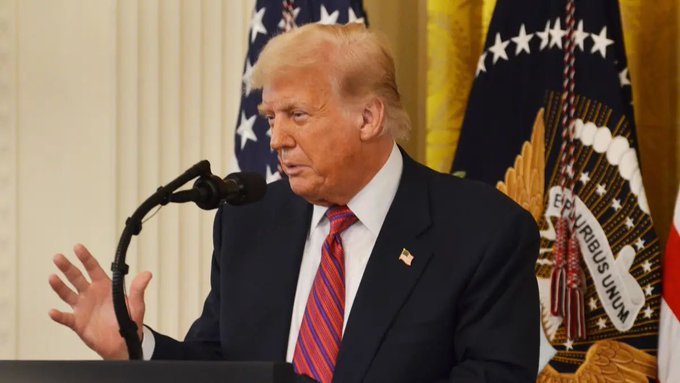 Donald Trump speaks at a podium, gesturing with his hand, with an American flag and presidential seal flag visible behind him.