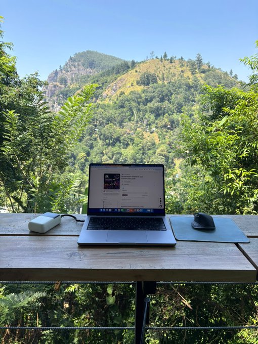 A laptop sits open on a wooden outdoor table next to a wireless mouse and charger, with a scenic view of a forested mountain and clear blue sky in the background.