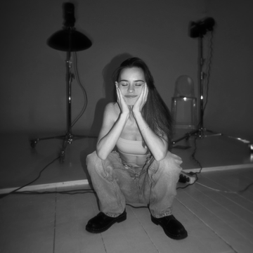 A black-and-white photograph of a young woman crouching on the floor in a studio setting, holding her face with both hands, with lighting equipment visible in the background.