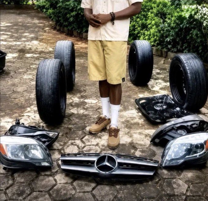 The image features a man standing next to several tires, including black and silver ones. He appears to be inspecting or working with these tires. The photo also includes a car part labeled "Mercedes" and another car part labeled "Day 22 Of June $SUI".