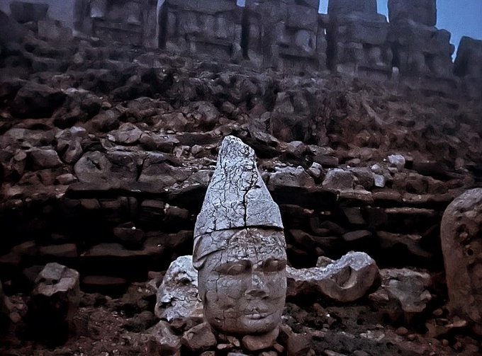 An archaeological site showing a large weathered stone head sculpture wearing a pointed headdress, partially buried in rubble at the base of ancient stone steps or ruins.