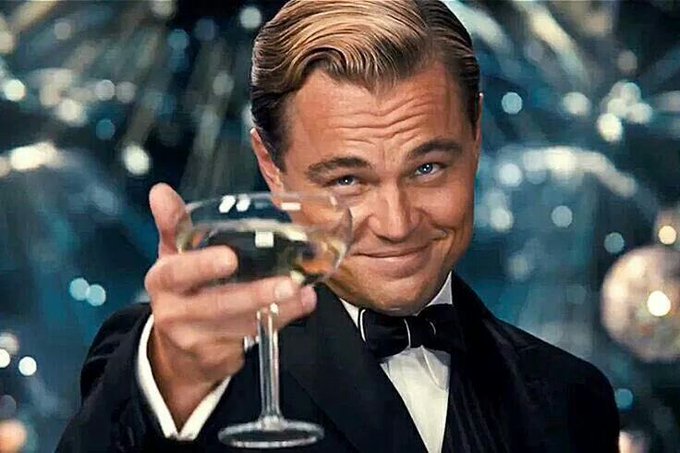 Leonardo DiCaprio as Jay Gatsby in a tuxedo, raising a champagne glass with a smile at a glittering party.