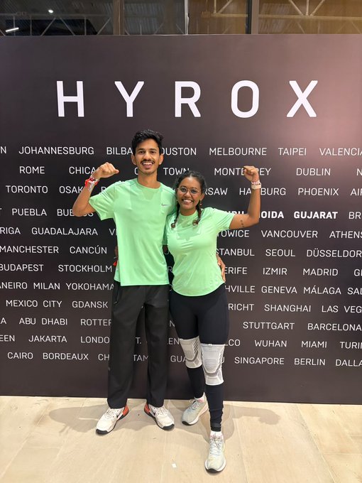 Two people in green t-shirts pose flexing their biceps in front of a HYROX backdrop listing various global cities including Johannesburg, Houston, Melbourne, and Taipei.