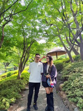 A young man and woman pose together on a tree-lined path, with the man saluting and the woman carrying a colorful bag.