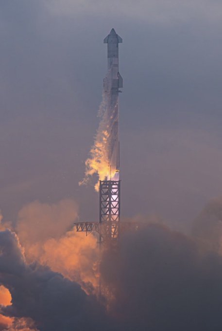 A SpaceX Starship rocket ascends during a test flight, with flames and exhaust billowing from its base against a dusky sky.