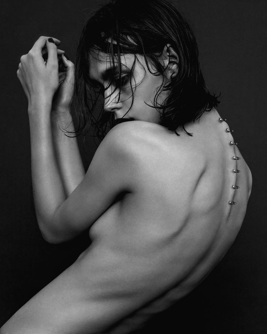 Black and white artistic photograph of a person with wet hair covering their face, posing with hands raised near their head and showing their bare back adorned with a decorative chain.