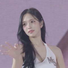 A young woman with long dark hair stands against a pink backdrop, wearing a white tank top and a delicate necklace.