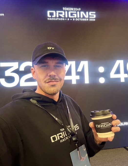 A man wearing a black hoodie and 'OV' cap holds a branded coffee cup at the TOKEN2049 ORIGINS hackathon event (4-5 October 2026), standing in front of a countdown timer display.