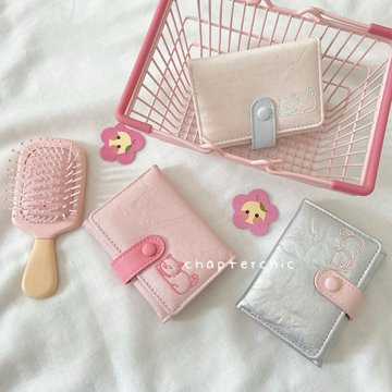A flat lay photograph featuring pastel pink and silver wallets, a pink hairbrush, and small koala decorations arranged on a white textured fabric with a pink wire basket.
