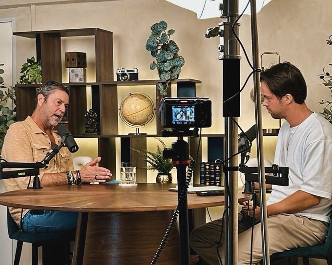 Two men sit across from each other at a round wooden table during what appears to be a podcast or interview recording session, with professional microphones, lighting equipment, and a camera on a tripod between them. The setting features decorative shelving with books, a globe, a vintage camera, and plants in the background, suggesting a polished studio environment for discussing bullish crypto market predictions and adoption cycles.