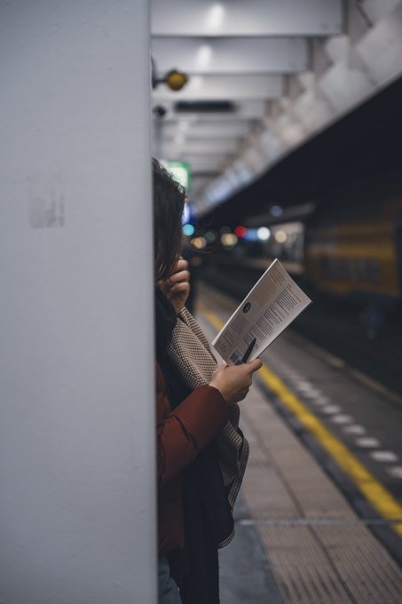 A woman stands on a train platform, partially hidden behind a pillar, reading a document while waiting near the yellow safety line.