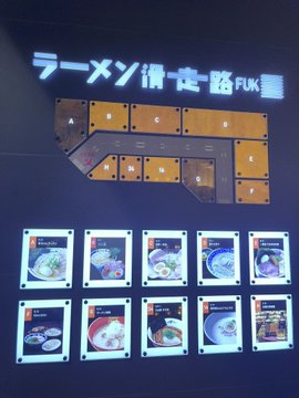 An illuminated menu display for a ramen restaurant called 'ラーメン博多一路 FUK' (Ramen Hakata Ichiro FUK) featuring photos of different ramen bowls on a black wall.