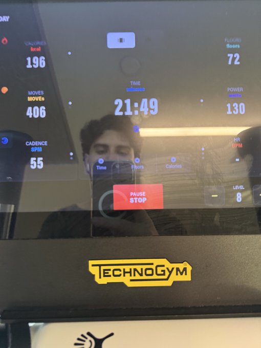 The image shows a person taking a selfie in front of a TechnoGym equipment display showing a workout in progress with 196 calories burned, 72 floors climbed, 406 moves, 21:49 elapsed time, 130 power, 55 cadence, and 8 level—which ties to the post by visualizing active exercise that contributes to the productivity and energy linked to earning more money.