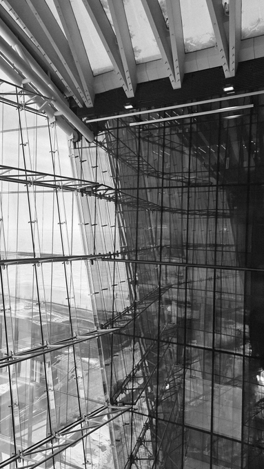 A black and white architectural photograph showing an interior view of a modern building with exposed steel beams, glass walls, and structural trusses.
