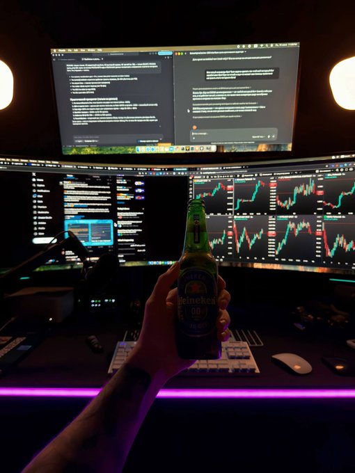 A hand holds a Heineken 0.0 bottle in front of a multi-monitor setup displaying trading charts and code, illuminated by purple underglow lighting.
